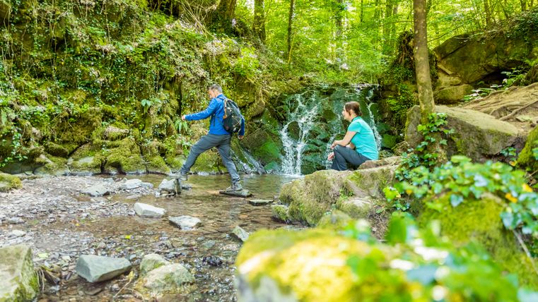 Zwei Personen in einem Wald, eine überquert einen Bach, die andere sitzt auf einem Felsen.