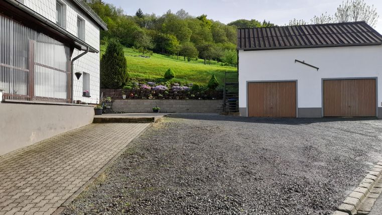 Eine ruhige Auffahrt mit einem Wohnhaus auf der linken Seite und einer Garage auf der rechten Seite. Im Hintergrund sind grüne Hügel und Bäume sichtbar.