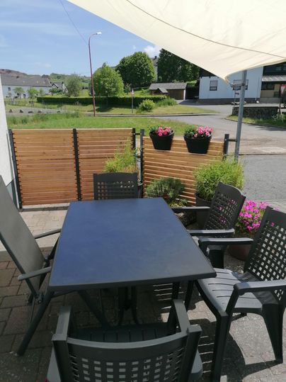 Ein gemütlicher Außenbereich mit einem schwarzen Tisch und Stühlen. Der Raum ist mit Pflanzen und blühenden Kästen dekoriert.