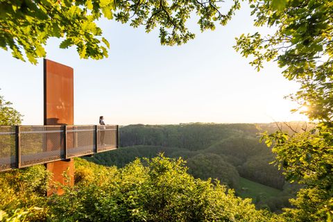 Person steht auf einem Skywalk mit Blick auf bewaldete Hügel bei Sonnenuntergang.