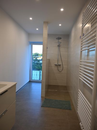Ein modernes Badezimmer mit einer Dusche und großen Fliesen. Ein Fenster sorgt für viel Tageslicht und einen Blick nach draußen.