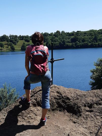 Eine Person sitzt auf einem Felsen mit Blick auf einen ruhigen See. Die Sonne scheint und die Umgebung ist grün und bewaldet.
