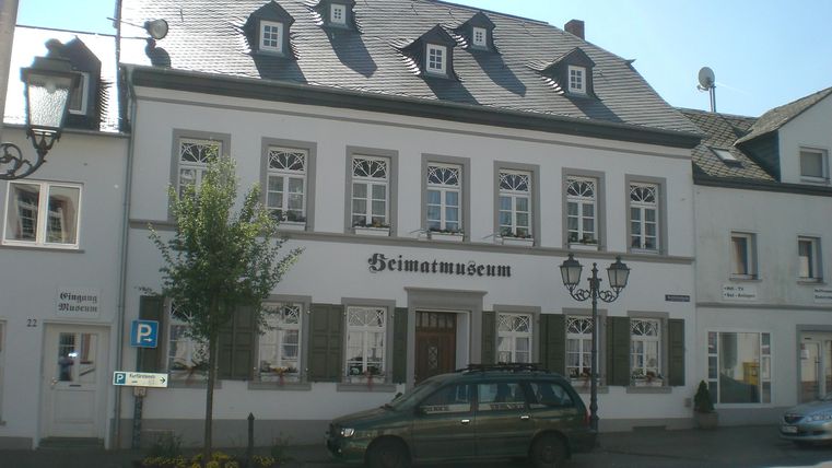 A historic building with a bright facade and a gabled roof. In front of the building is a car and there are some trees nearby.
