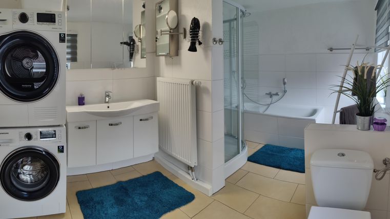Ein modernes Badezimmer mit Waschmaschine und Trockner, einem Waschbecken und einer Dusche. Es gibt blaue Matten und eine Fensterbank mit Pflanzen.