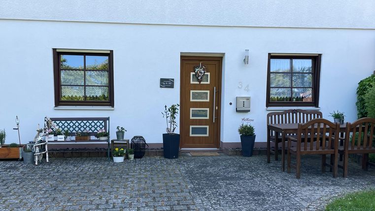 Ein einladender Hauseingang mit einer Holztür und zwei Fenstern. Vor der Tür befinden sich Blumentöpfe und eine Sitzbank.