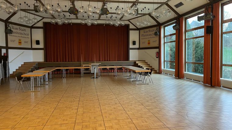 Ein großer Veranstaltungsraum mit Holzfußboden und viel Tageslicht. Die Wände sind mit Vorhängen und Fenstern ausgestattet, und Tische sind im Raum aufgestellt.