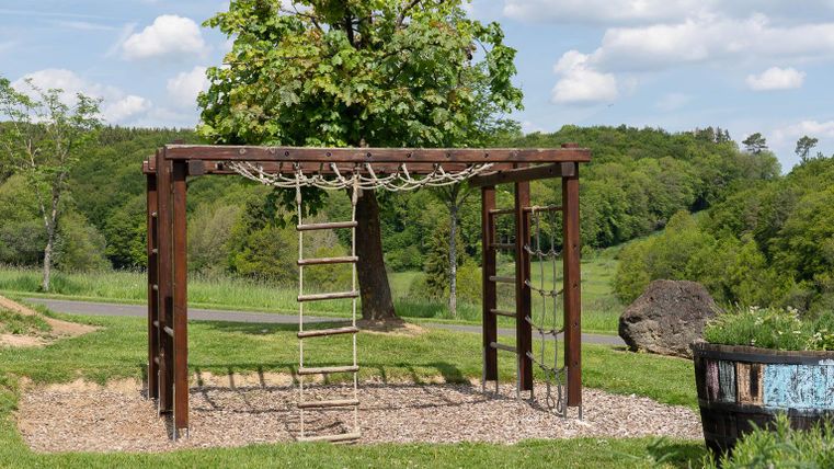 Ein Spielplatz mit einer Kletterapparate aus Holz und Seilen. Im Hintergrund sind grüne Bäume und ein blauer Himmel sichtbar.