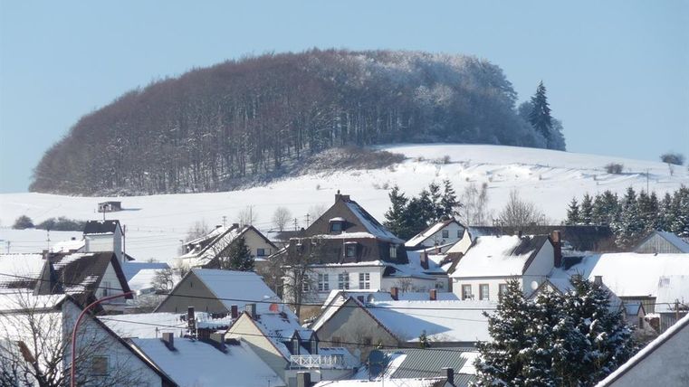 Eine schneebedeckte Landschaft mit einem Hügel im Hintergrund und einem kleinen Dorf im Vordergrund. Die Dächer der Häuser sind mit Schnee bedeckt und die Bäume am Hügel sind winterlich verschneit.