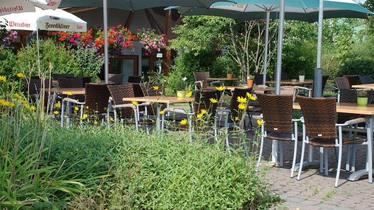 Ein gemütlicher Außenbereich mit Tischen und Stühlen unter Sonnenschirmen. Umgeben von bunten Blumen und grüner Vegetation.