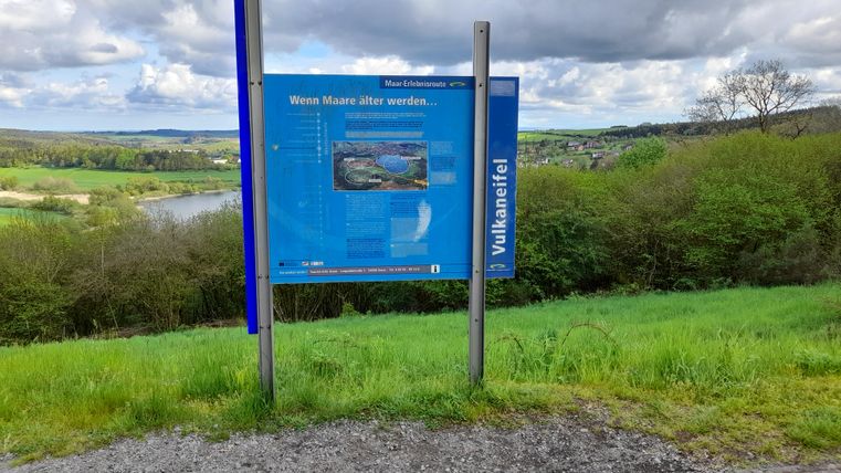 Ein Informationsschild am Hang mit Blick auf eine grüne Landschaft und einen See. Der Himmel ist bewölkt und die Umgebung ist ruhig und naturnah.