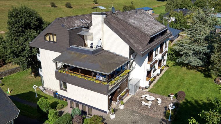 Ein großes Haus mit mehreren Etagen und einem Balkon. Der Garten ist gut gepflegt und umgeben von grünen Feldern.