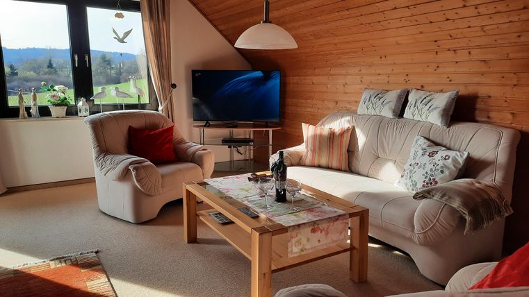 Ein gemütliches Wohnzimmer mit einer Holzvertäfelung und großen Fenstern. Es gibt bequeme Sofas, einen Couchtisch und einen Fernseher.