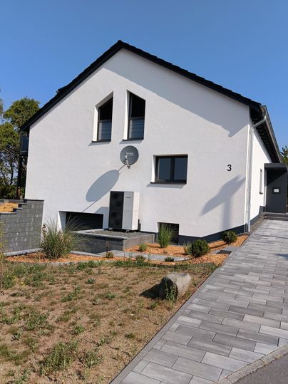 A modern, white house with an appealing design. The front yard is well-maintained and designed with stones and plants.