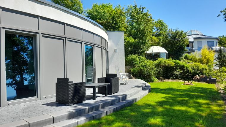 Ein modernes Haus mit großen Fenstern und einem gepflegten Garten. Auf der Terrasse stehen schwarze Gartenmöbel und es ist sonnig.