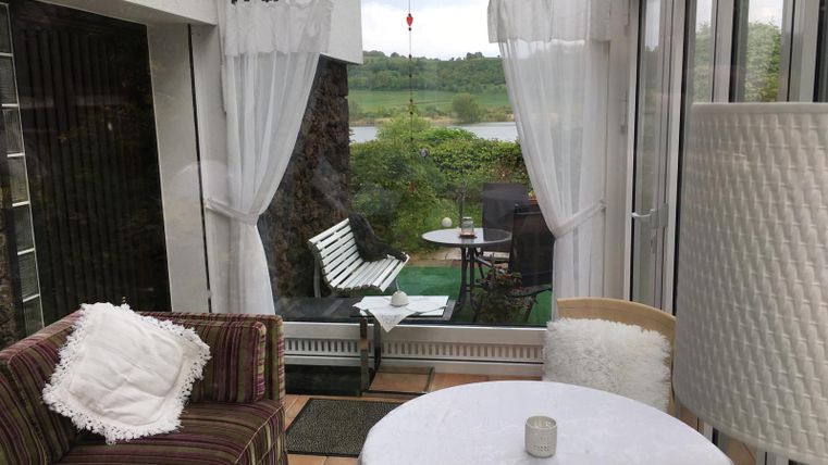 Eine gemütliche Sitzecke in einem Wintergarten mit Blick ins Grüne. Helle Vorhänge lassen viel Licht herein und schaffen eine einladende Atmosphäre.