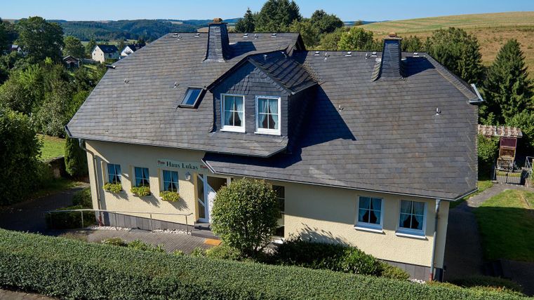 Ein hübsches Haus mit einem Steildach und großen Fenstern. Umgeben von grünen Büschen und einer ländlichen Landschaft.