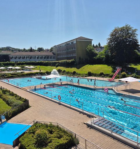 Ein großes Schwimmbad mit mehreren Becken und einer Rutsche. Im Hintergrund sieht man Sonnenschirme und eine grüne Rasenfläche.