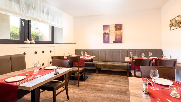 Ein elegantes Restaurant mit Holztischen und roten Tischdecken. Die Wände sind dekoriert und das Licht schafft eine angenehme Atmosphäre.