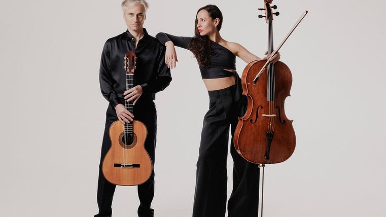Ein Musiker mit einer Gitarre steht neben einer Musikerin mit einem Cello. Beide tragen elegante schwarze Kleidung und posieren vor einem neutralen Hintergrund.