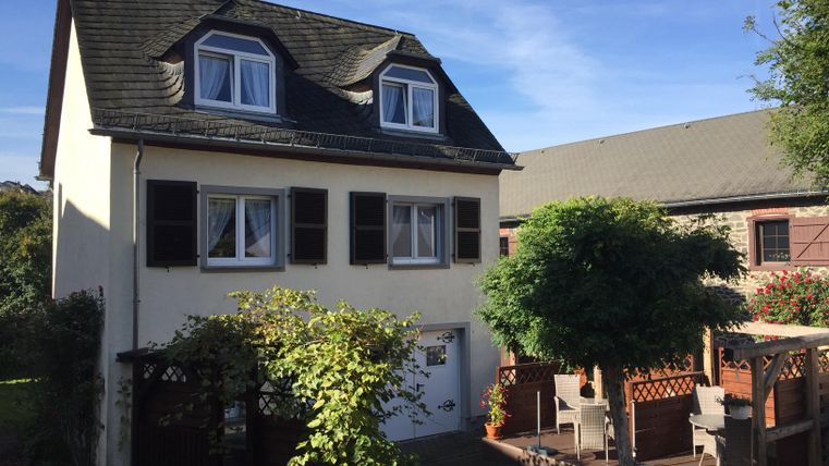 Ein zweigeschossiges Haus mit weißer Fassade und braunen Fensterläden. Im Vordergrund befindet sich ein Garten mit Möbeln und einem kleinen Baum.