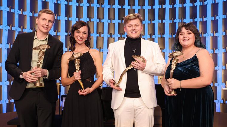 Vier personen staan in elegante kleding en houden elk een onderscheiding in de hand. Op de achtergrond is een stijlvolle podiumdecoratie te zien.
