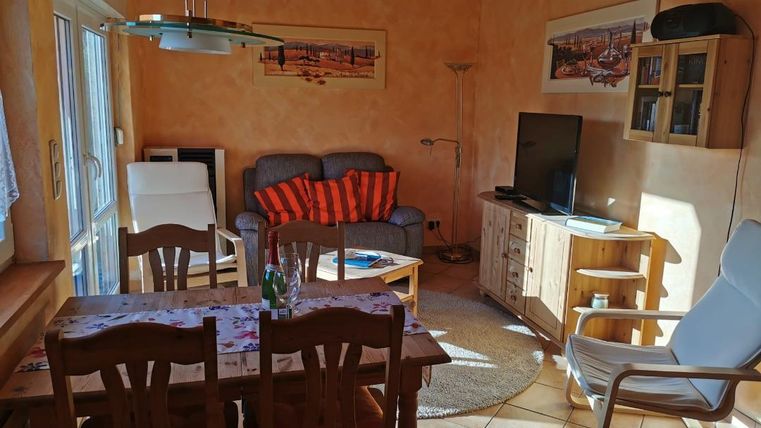 Ein gemütliches Wohnzimmer mit einem Esstisch und Stühlen. Auf der Couch liegen orangefarbene Kissen und ein Fernseher steht auf einem Holzschrank.