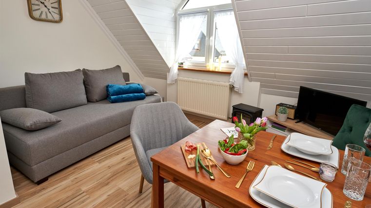 Ein gemütliches Zimmer mit einem grauen Sofa und einem Esstisch. Der Tisch ist schön gedeckt und ein Fenster sorgt für viel Tageslicht.