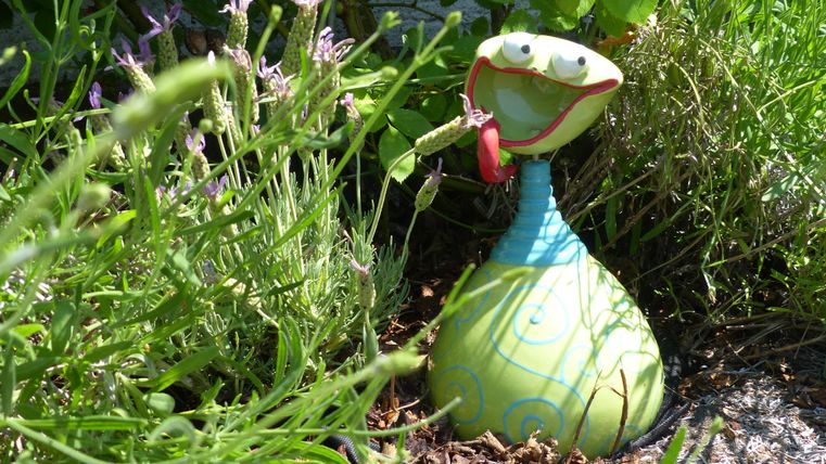 Eine bunte Gartenfigur in Form einer Schlange steht zwischen grünem Gras und bunten Blumen. Sie hat ein fröhliches Gesicht und hält etwas in der Hand.