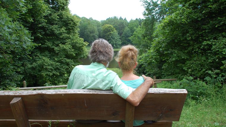 An older couple is sitting on a bench in the greenery.
They enjoy the peace and the view of nature.