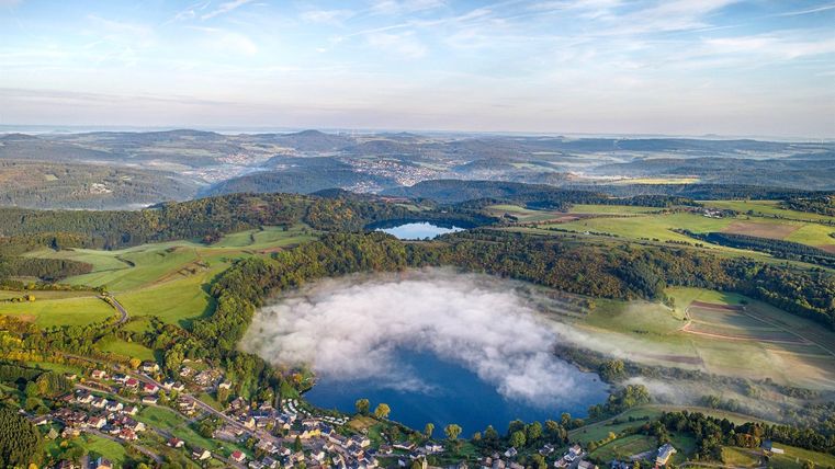 Eine malerische Landschaft mit einem See und Nebel über dem Wasser. Umgeben von grünen Feldern und kleinen Dörfern.