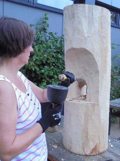 Eine Frau schnitzt an einem Holzblock im Freien. Sie verwendet ein Werkzeug, um eine Öffnung in das Holz zu schaffen.