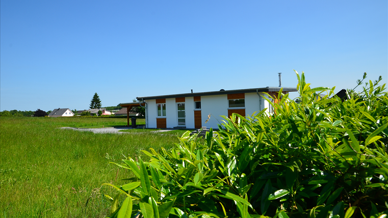 Ein modernes Haus steht inmitten einer grünen Wiese unter klarem, blauem Himmel. Im Hintergrund sind weitere Häuser sichtbar.