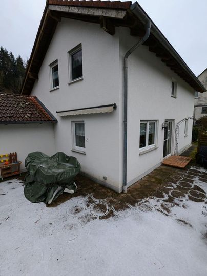 Ein zweistöckiges, weißes Gebäude mit einem Stegdach. Im Vordergrund liegt ein gepflasterter Bereich mit einem abgedeckten Objekt.
