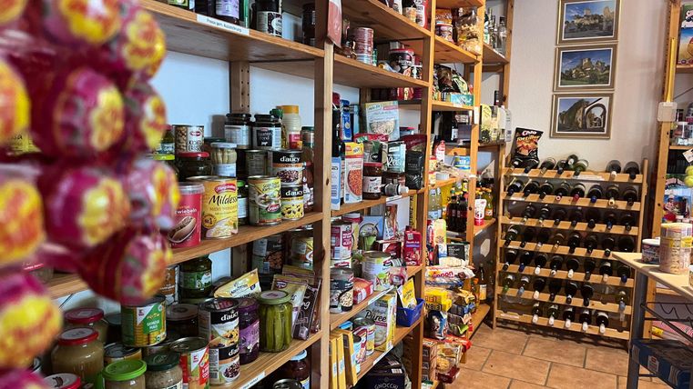 Ein gut sortierter Lebensmittelladen mit Regalen voller Gläser, Dosen und Snacks. Im Hintergrund sind auch Weinfässer zu sehen.