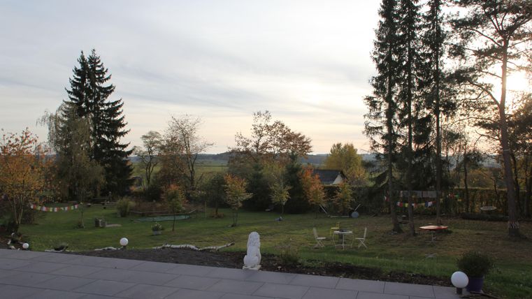 Eine ruhige Gartenlandschaft mit herbstlichen Bäumen und einem klaren Himmel. Im Vordergrund sind Sitzgelegenheiten und einige Gartenornamente sichtbar.