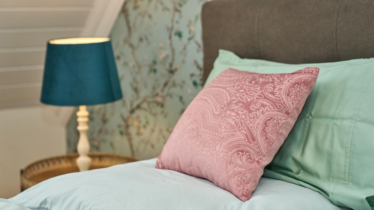 Ein gemütliches Schlafzimmer mit einem hellblauen Bett und einem rosafarbenen Kissen. Eine Lampe mit grünem Shade sorgt für sanftes Licht.