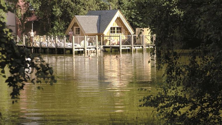 Ein gemütliches Haus am Wasser mit einer Holzterrasse. Umgeben von Bäumen spiegelt sich das Gebäude im ruhigen See.