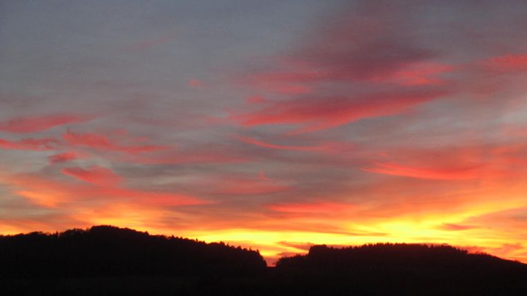 Ein atemberaubender Sonnenuntergang mit leuchtenden roten und orangefarbenen Wolken. Silhouetten von Hügeln sind im Vordergrund sichtbar.