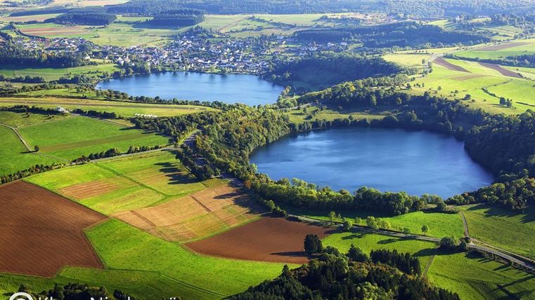 Eine Luftaufnahme von zwei Seen umgeben von grünen Wiesen und Feldern. Im Hintergrund ist ein Dorf sichtbar, das von der schönen Landschaft umgeben ist.