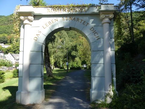 Ein eleganter Torbogen in einem parkähnlichen Umfeld. Der Weg führt durch grüne Bäume und Pflanzen.