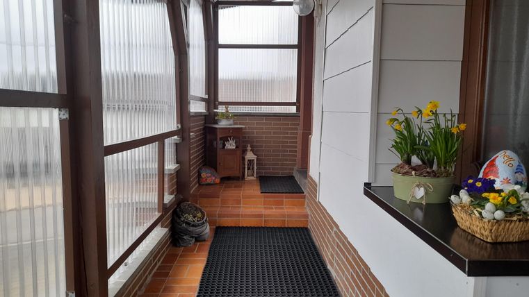Ein heller Flur mit Glaswänden und braunen Fliesen. Auf der Fensterbank stehen frische Blumen und ein Osternest.