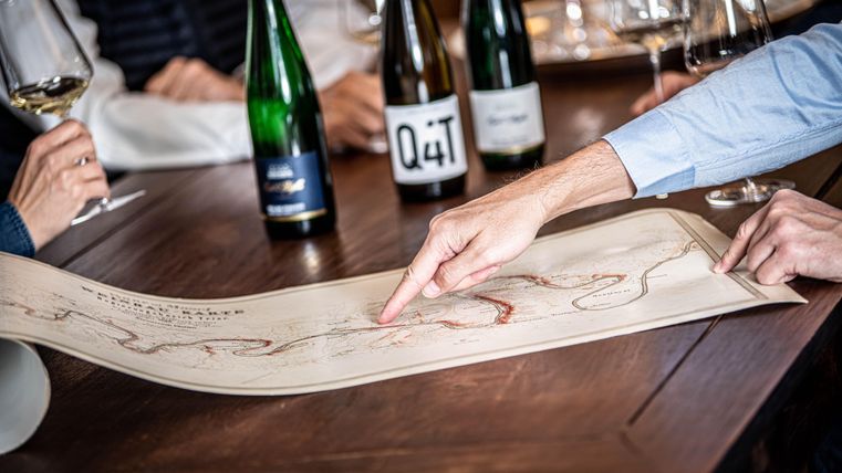 Eine Gruppe von Menschen sieht sich eine große Karte an und zeigt auf einen bestimmten Punkt. Im Hintergrund stehen einige Flaschen Wein und Gläser auf einem Tisch.