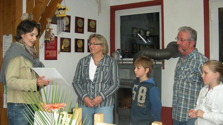 Eine kleine Gruppe von vier Personen steht in einem gemütlichen Raum. Eine Frau liest ein Dokument vor, während die anderen aufmerksam zuhören.