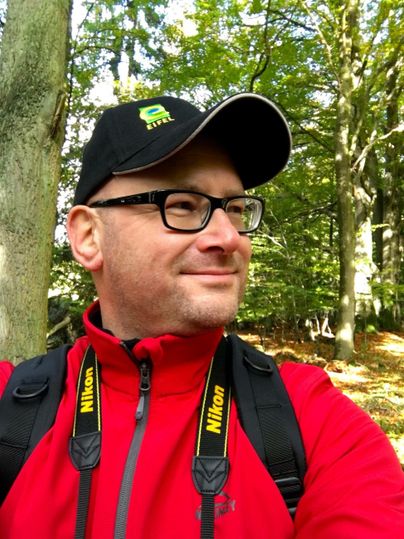 Ein Mann in einem roten Jackett steht in einem Wald. Er trägt eine Mütze und eine Kamera um den Hals.