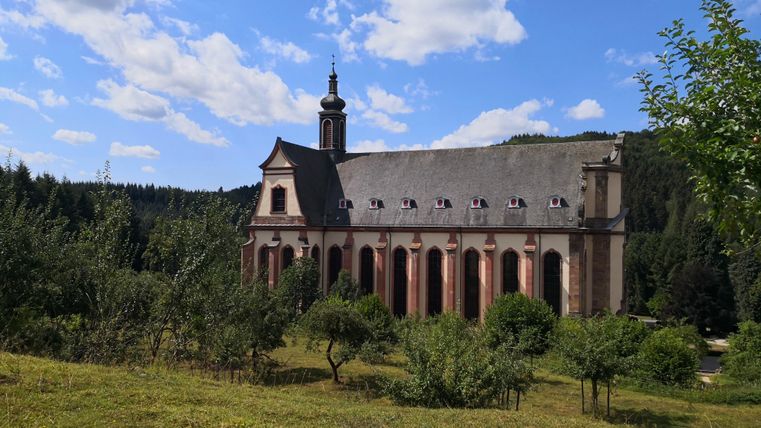 Eine beeindruckende Kirche umgeben von Bäumen und Wiesen. Der Himmel ist klar und blau, mit einigen Wolken.