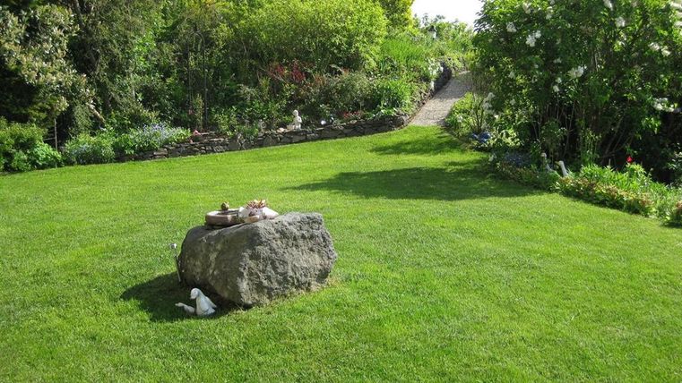 Ein gepflegter Garten mit grünem Rasen und einem großen Stein in der Mitte. Im Hintergrund sind bunte Sträucher und ein Weg sichtbar.