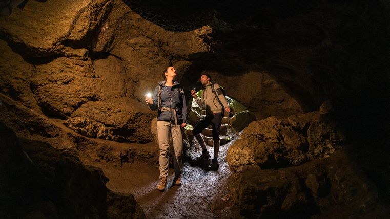 Zwei Personen in Wanderkleidung erkunden eine Höhle mit Taschenlampen. Die Umgebung ist dunkel und felsig.