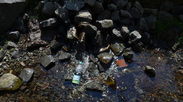 Ein kleiner Teich mit klaren Wasser und Steinen. Einige Flaschen liegen im Wasser.