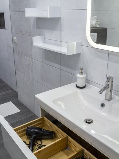 Ein modernes Badezimmer mit einer weißen Waschbecken- und Regalkombination. Im offenen Schubladen liegen ein Haartrockner und andere Badutensilien.