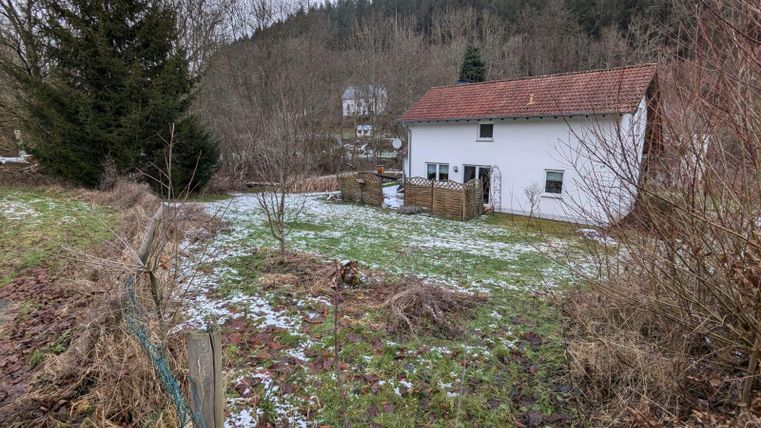 Ein einfaches Haus in einer ländlichen Gegend, umgeben von Bäumen und Winterlandschaft. Der Boden ist teilweise mit Schnee bedeckt.